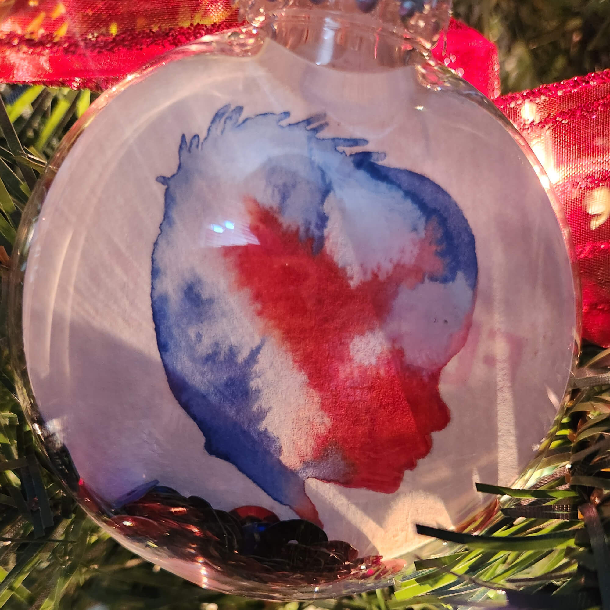 Red, White and Blue Silhouette in a 3.15" clear, Shatterproof ornament on an evergreen background with lights and red ribbon