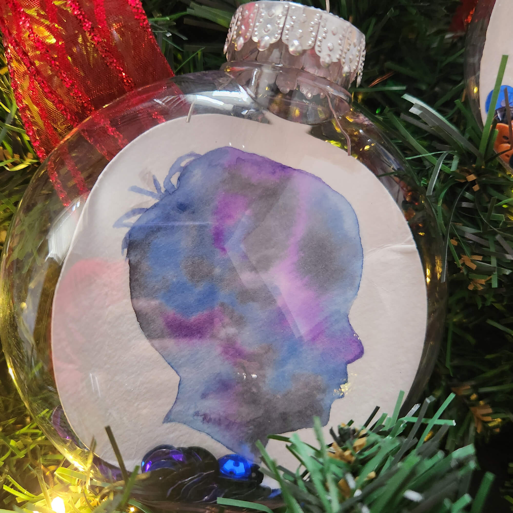 Purple, Blue and Black Silhouette in 3.94" Clear Shatterproof Ornament in an evergreen background with lights and ribbon